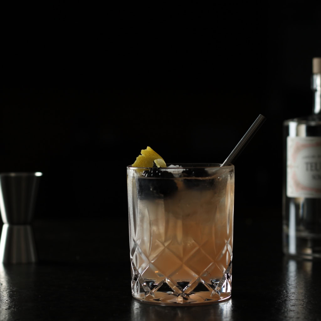 Bramble Cocktail im facettierten Glas mit Eis, Zitronenzeste, Brombeeren und Metallstrohhalm