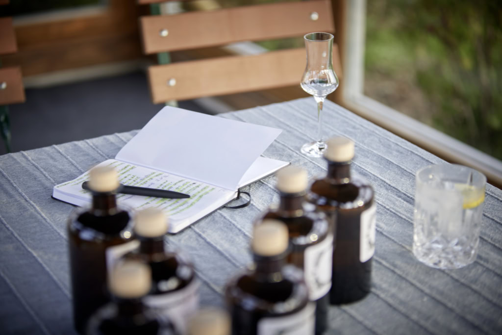 Mehrere braune Probeflaschen mit hellen Verschlüssen stehen auf einem Tisch neben einem Notizblock, einem Stift und einem Verkostungsglas in einem hellen Raum.