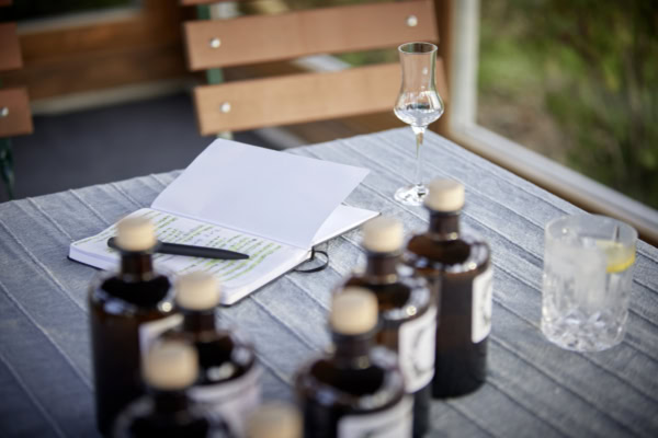 Mehrere braune Probeflaschen mit hellen Verschlüssen stehen auf einem Tisch neben einem Notizblock, einem Stift und einem Verkostungsglas in einem hellen Raum.